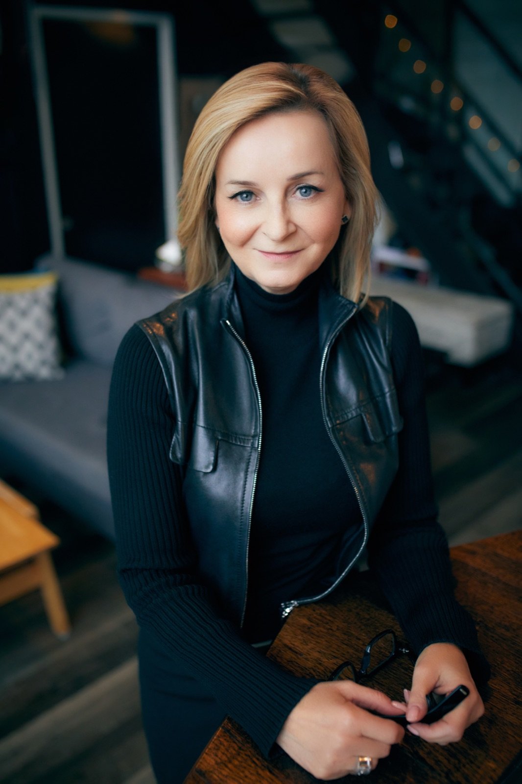 Tracy Quinton leaning on a walnut table in a black leather jacket, looking directly at the camera