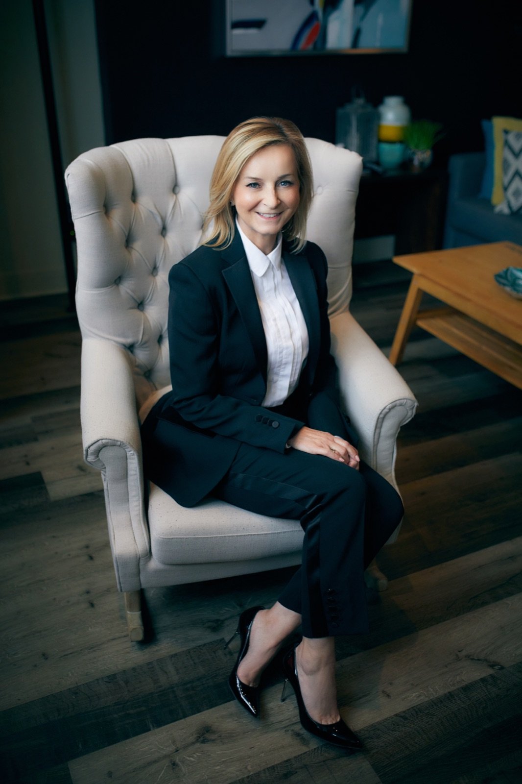 Tracy Quinton sitting in a tufted armchair in a navy suit, full length